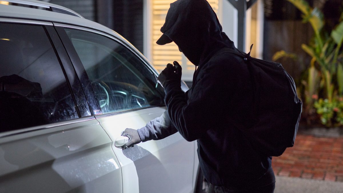 A person clad in all-black looks to open a car door at night. They are shining a torch on the handle and looking suspicious