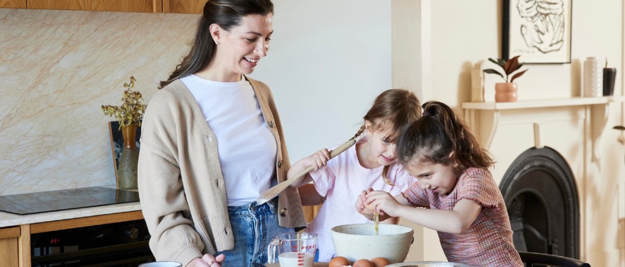 A mother and her two young daughters happily cooking at kitchen table. An induction cooktop can be seen in the background.