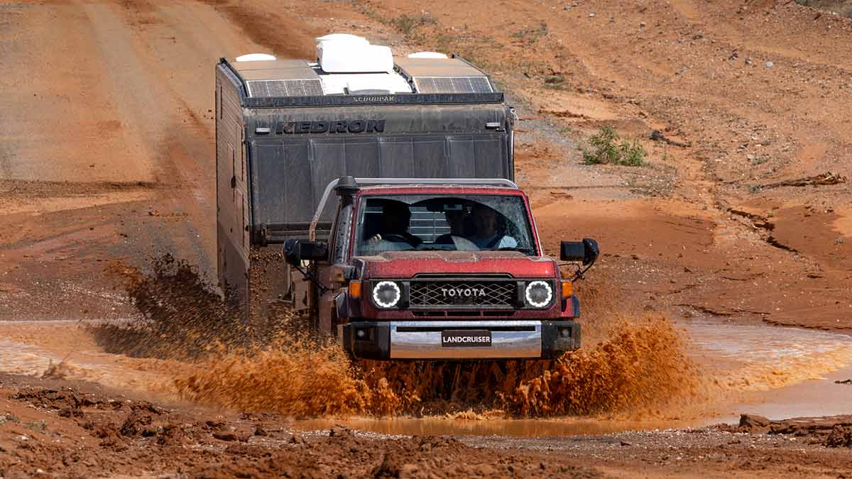 Red Toyota LandCruiser 70 Series GXL ute with caravan driving through muddy water crossing in barren outback location. 