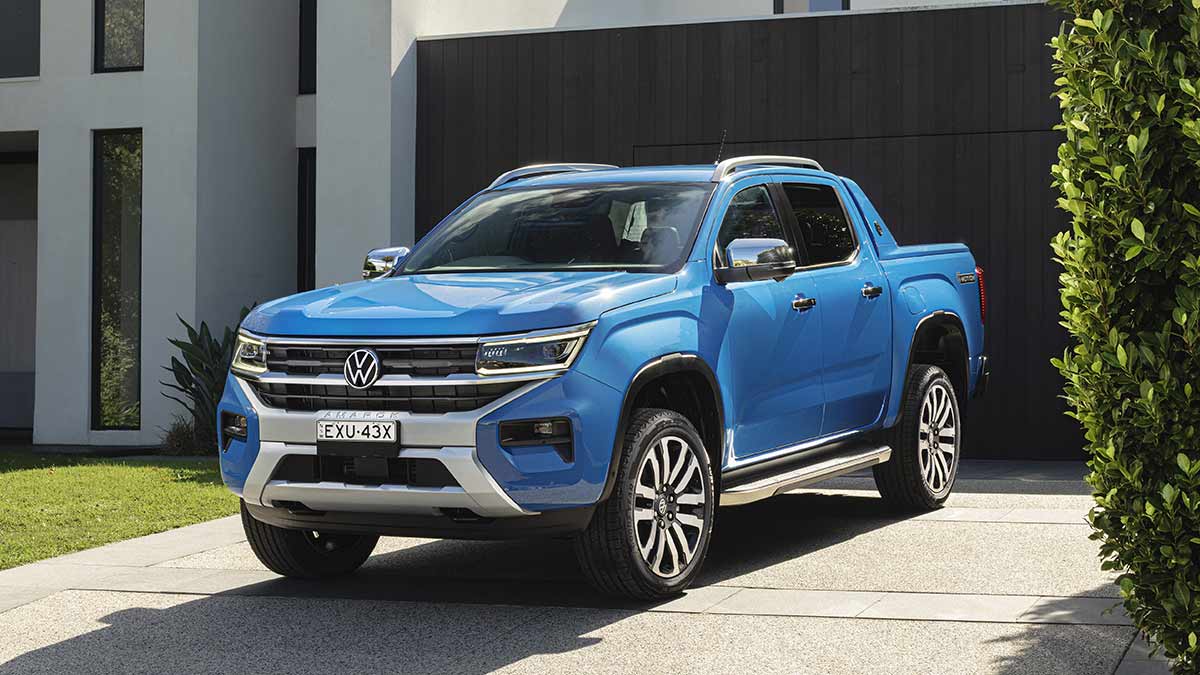 Blue Volkswagen Amarok Aventura dual-cab ute parked on driveway outside modern white house with tall dark garage door.