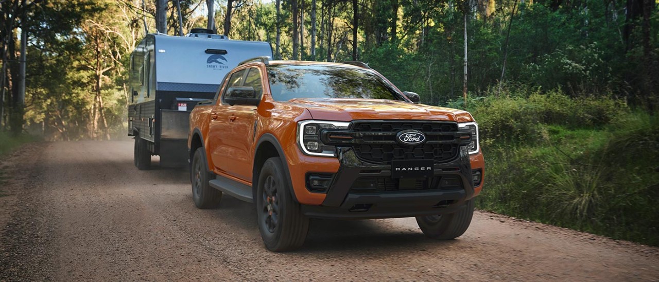 Orange Ford Ranger Wildtrak dual-cab ute towing caravan on shadowy treelined scrubby gravel road.