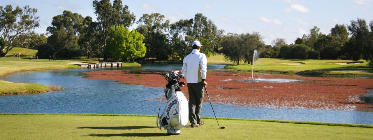 A man with a goldf caddy standing in front of a glittering pond on a golf course