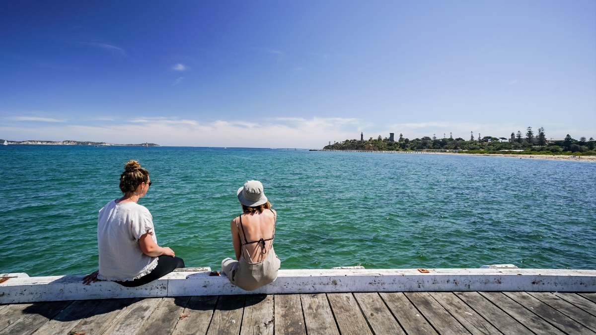 two women sitting on pier by ocean looking at headland