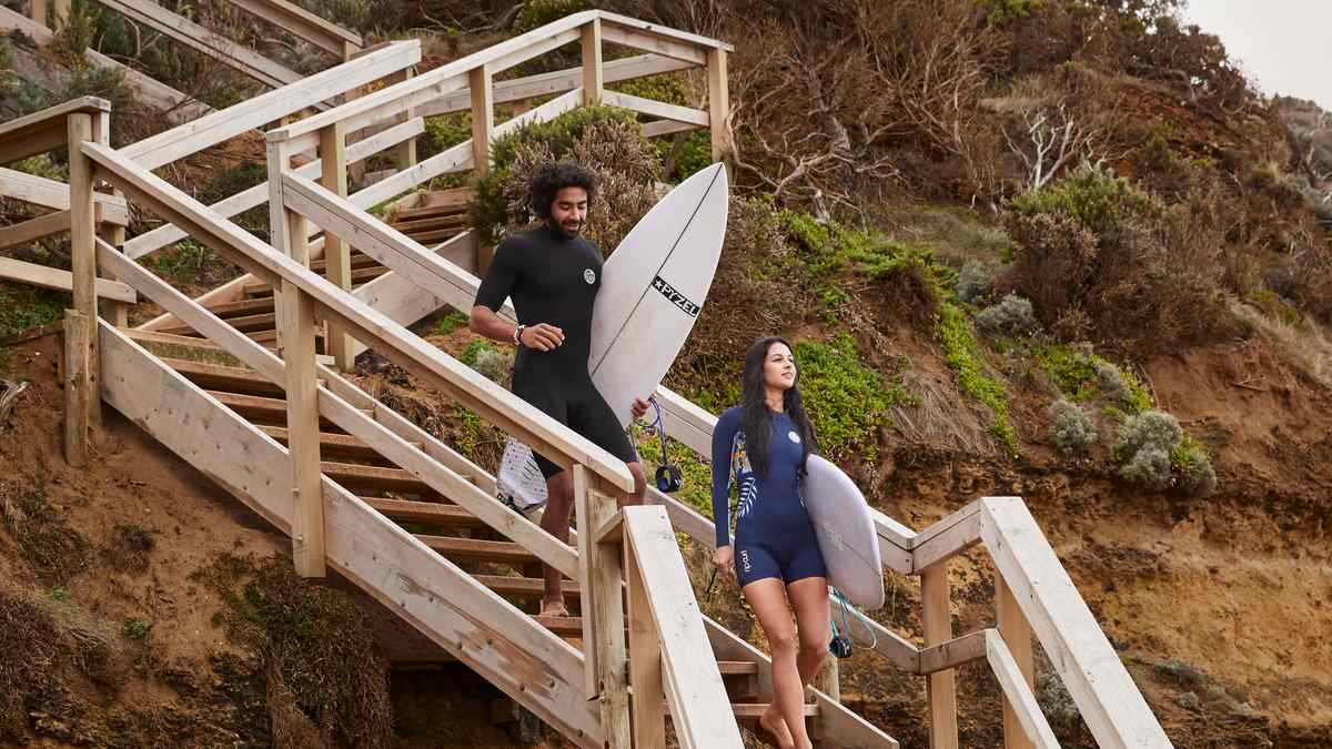 A man and woman both wearing short wetsuits and carrying surfboards walk down steps to Torquay Surf Beach