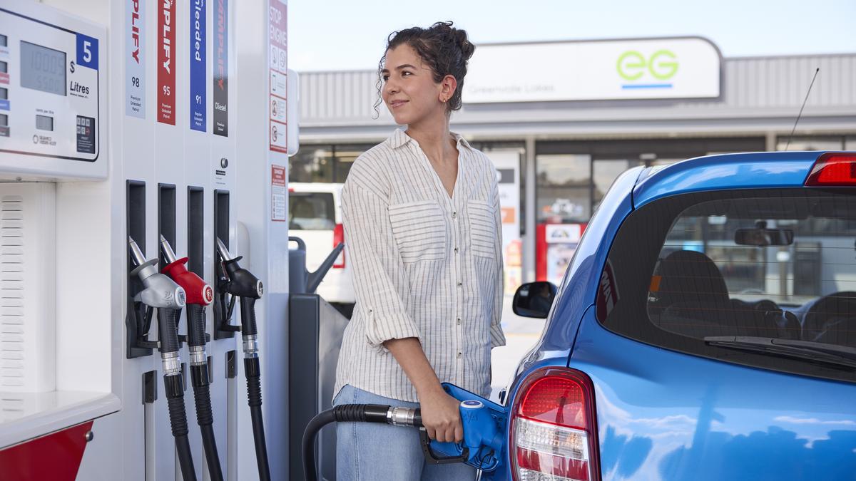 A woman filling up her blue Nissan Micra at a petrol bowser. She is smiling as she fills up her car
