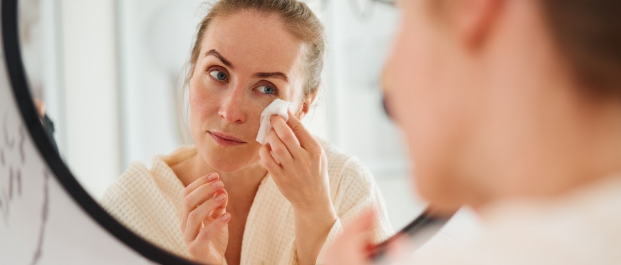 A woman looking in a round mirror while cleaning her face with a wet wipe
