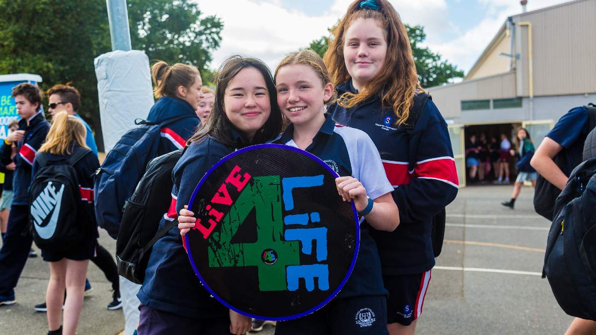 Three young women in school uniform smiling as they hold up a circular 'Live4Life' sign