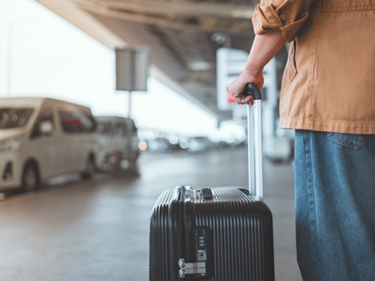 Rear view of male traveler standing with suitcase waiting for taxi at outside airport.