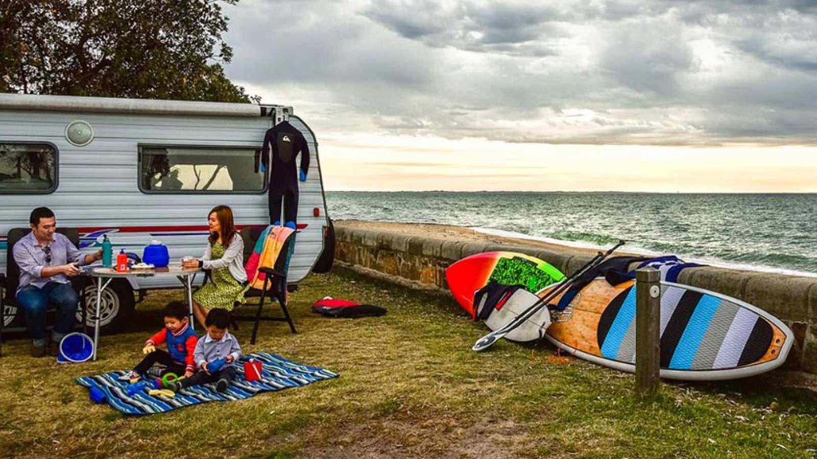 Young family sitting outside of a caravan next to the beach.