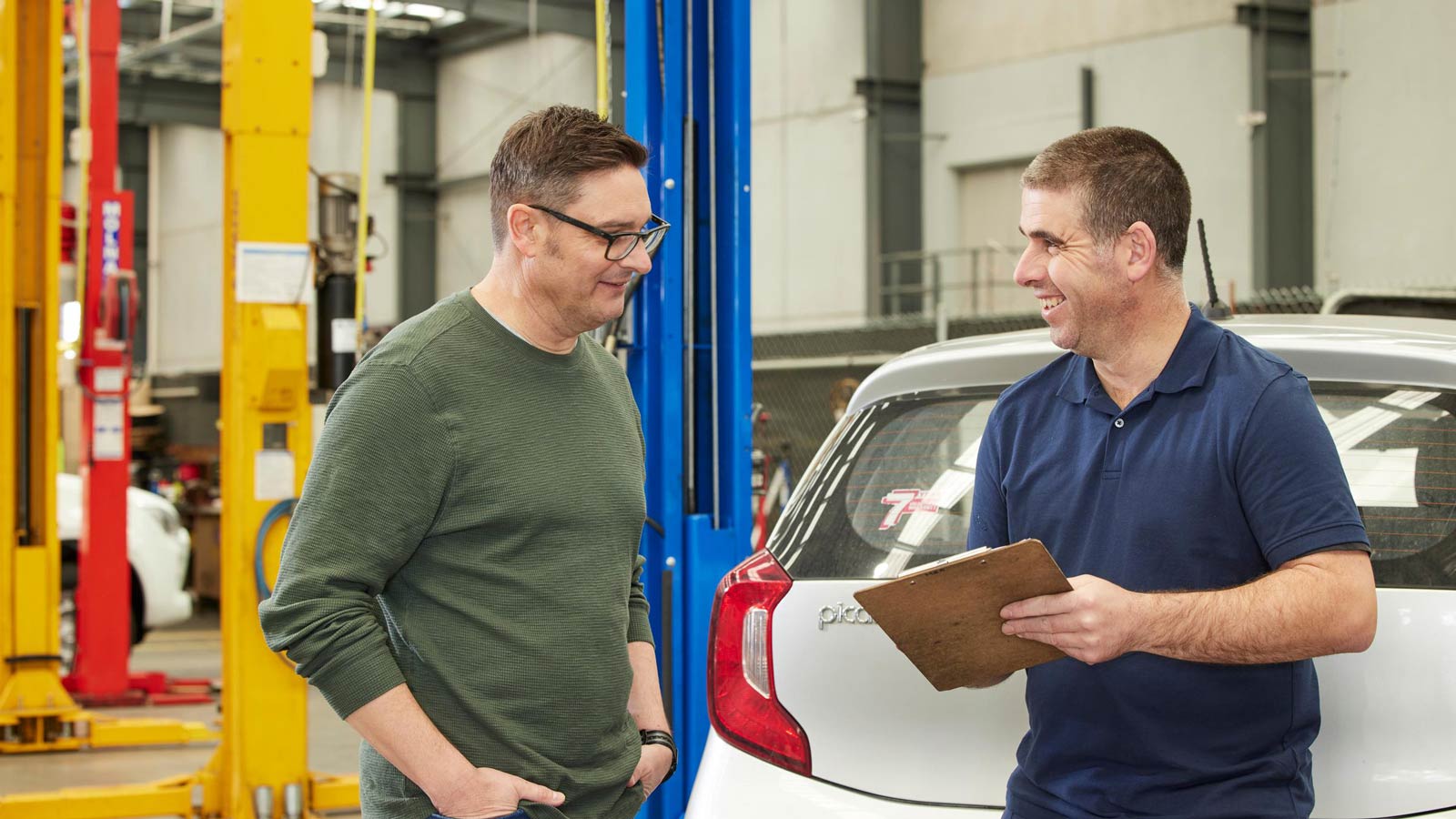 Mechanic with a clipboard smiling at a customer in a workshop.