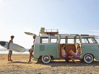 Young people with surfboards loading a blue combi van parked on the beach.