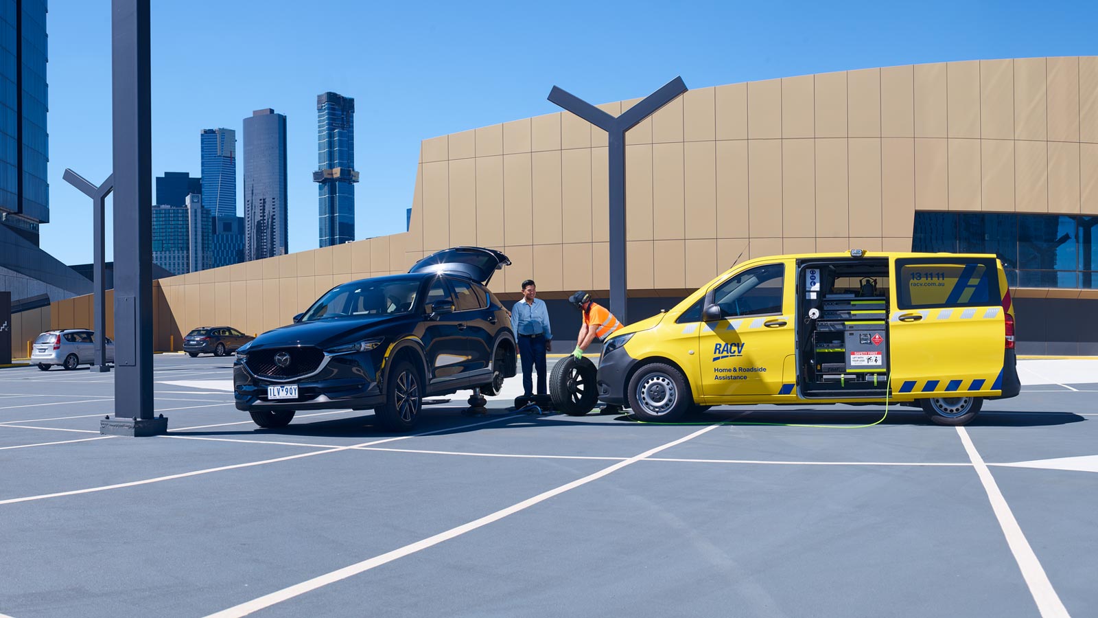 RACV roadside assistance mechanic replacing a car's tyre while speaking with a smiling customer.
