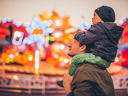 Child sitting on father's shoulders at a carnival, with red and yellow lights out of focus in the background.