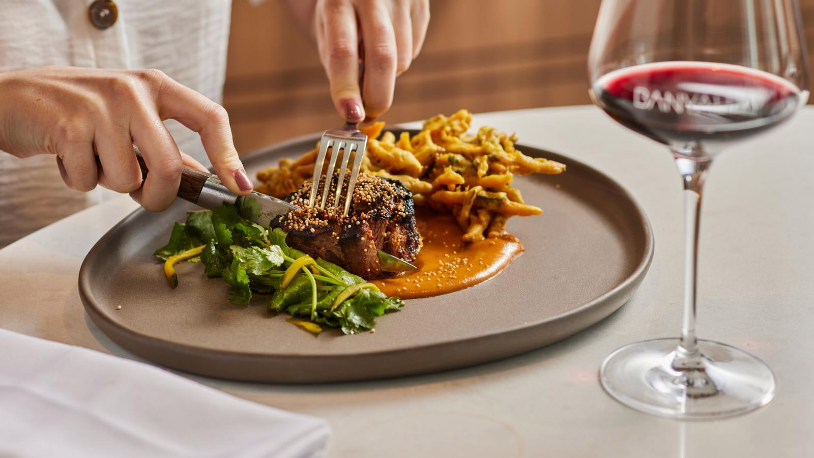 Person cutting through meat on plated dish with red wine glass on the side at Banyalla restaurant at RACV Healesville Country Club and Resort.