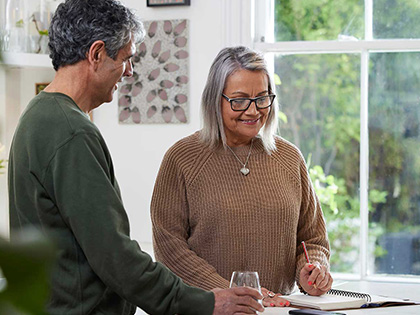 An older woman with grey hair and glasses stands in the kitchen in front of a bench, in front of her is an open notebook and a she is smiling. A smiling man with greying hair stands next to her, holding a glass of water, also smiling. 