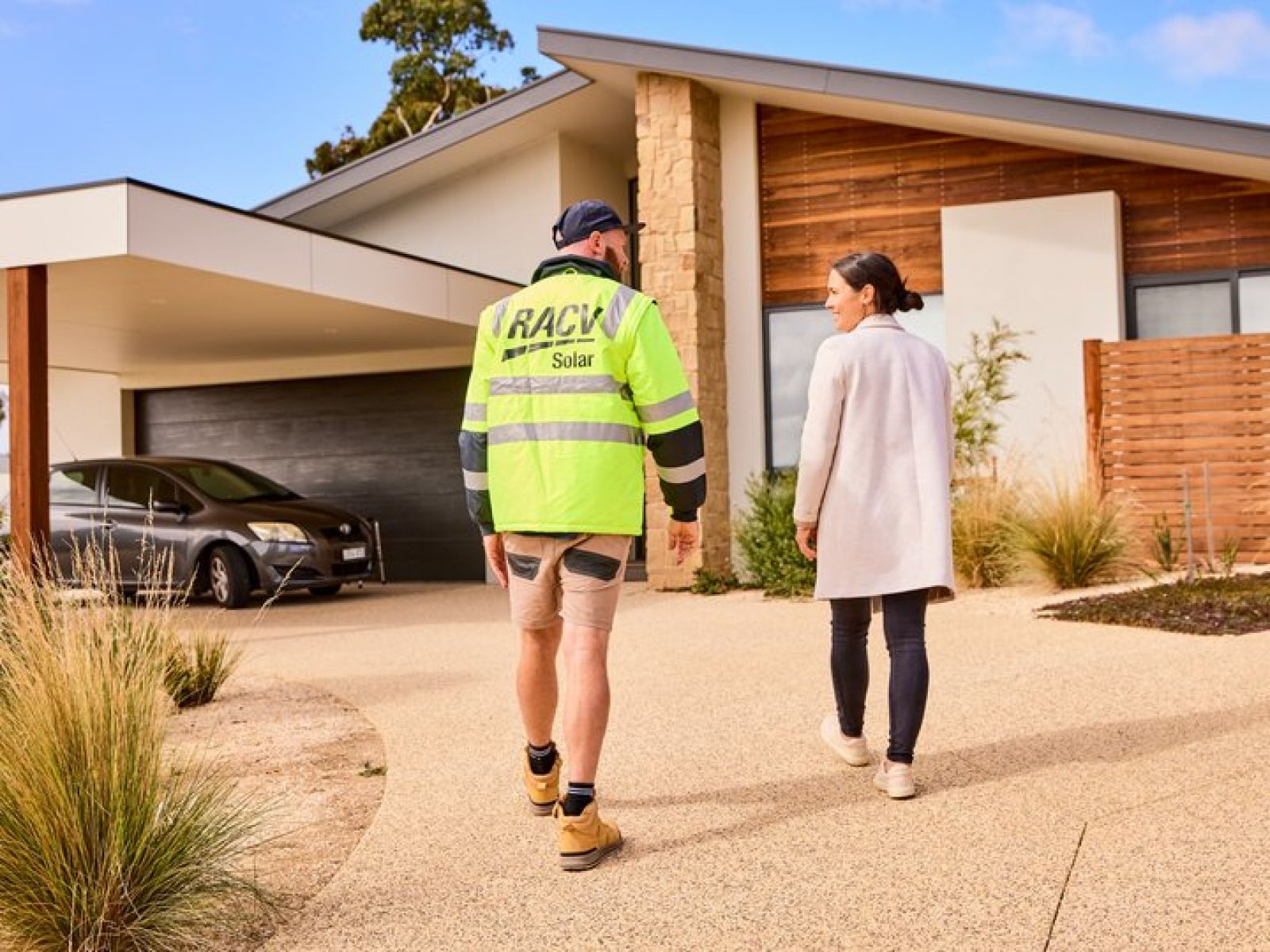 RACV Solar installer walking with a customer towards the entrance of their house, with a black car parked in the carport nearby.