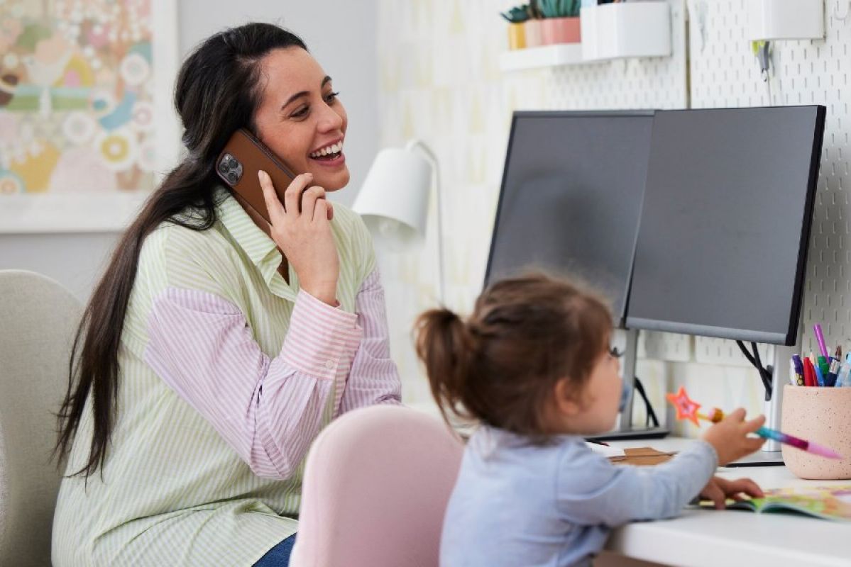 Woman on phone smiling in front of a monitor with her daughter drawing next to her