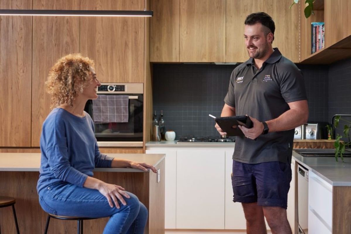 A man smiles while holding a tablet device like a checklist. A woman sits at a barstool in a home kitchen next to him smiling back