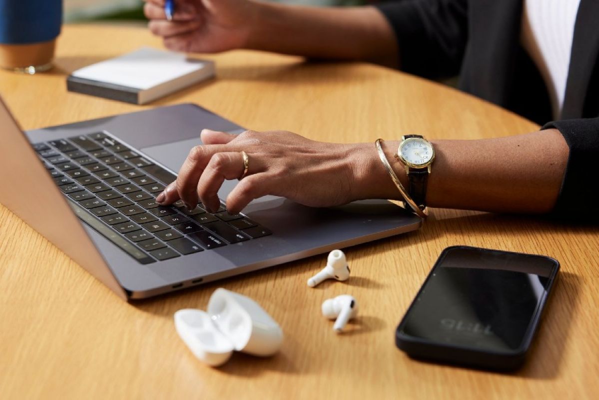 A close-up of a person working on a laptop. They're wearing a gold bangle, ring and watch and have a pair of earbuds  and a smartphone next to them on their desk