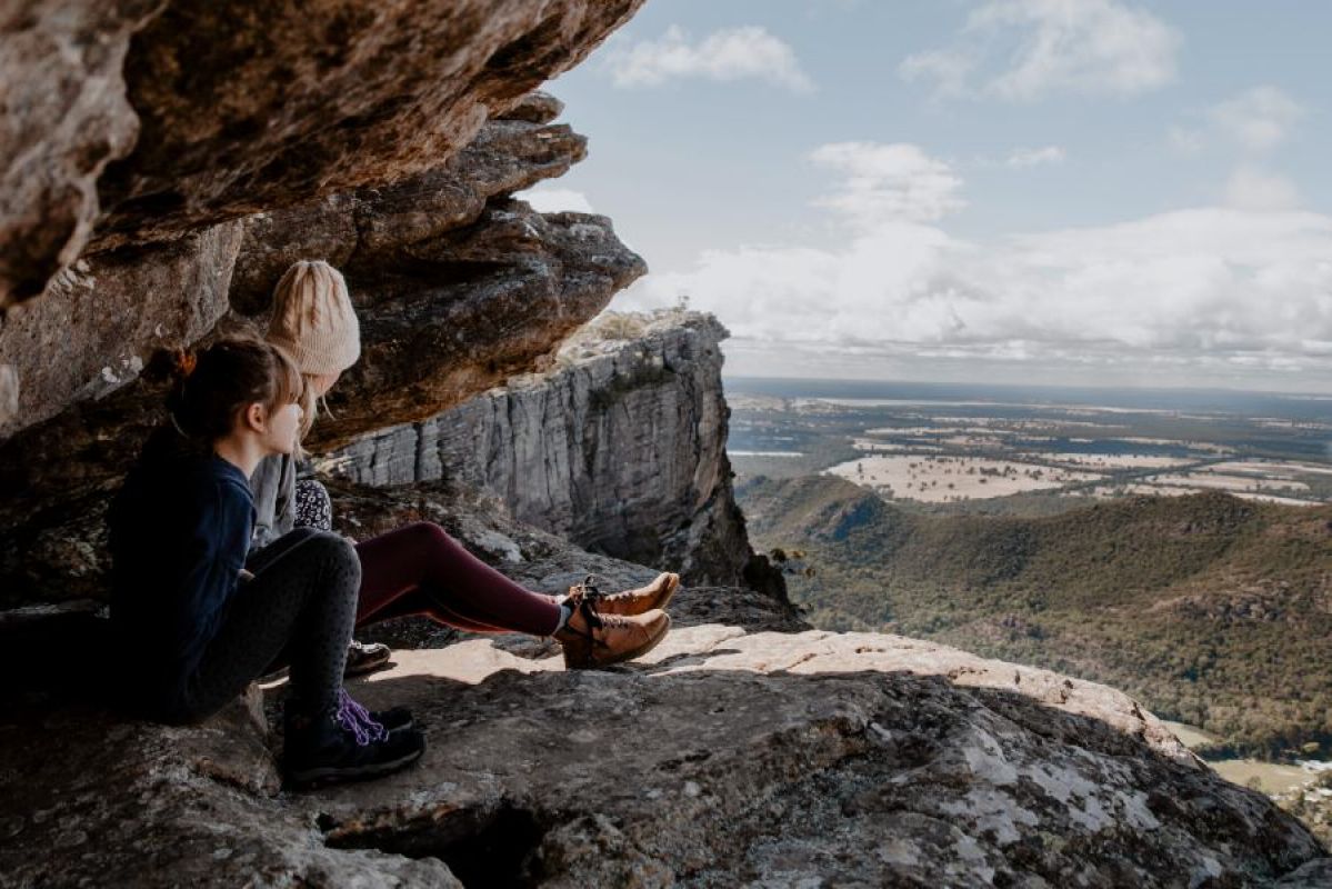 Two people sitting on a rock ledge in the GRampians and overlooking a spectacular vista of the land below