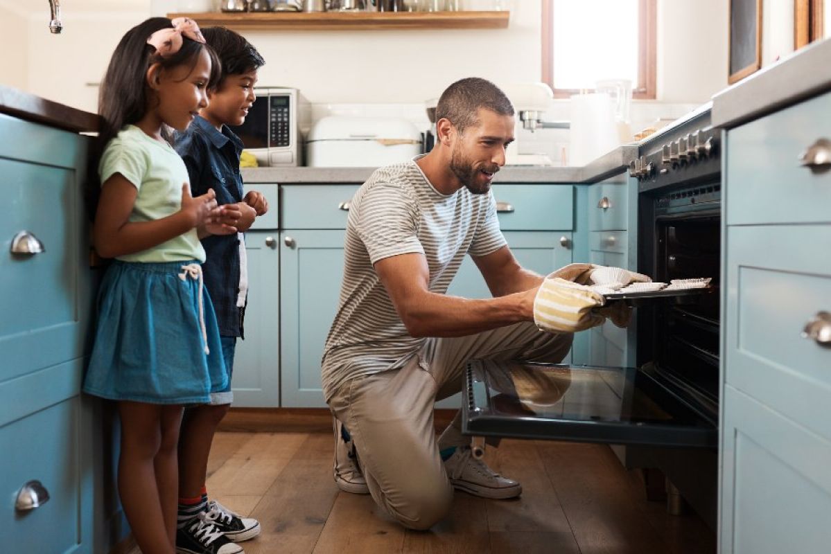 A man putting food into an oven while his two young children watch on happily