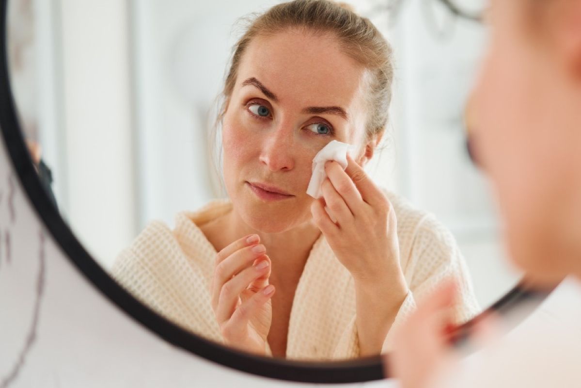 A woman looking in a round mirror while cleaning her face with a wet wipe