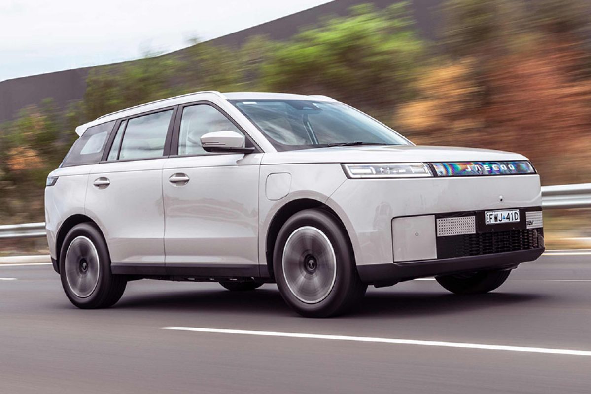 White Jaecoo J5 EV small SUV on freeway in left lane next to armco, small trees and tall grey sound barrier.