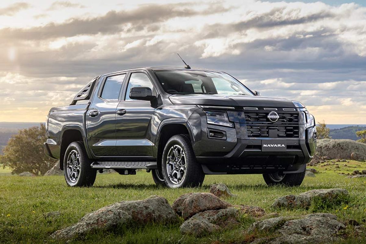Black Nissan Navara ST dual cab ute on grassy, rocky hilltop in rural area with bright blue sky and clouds.