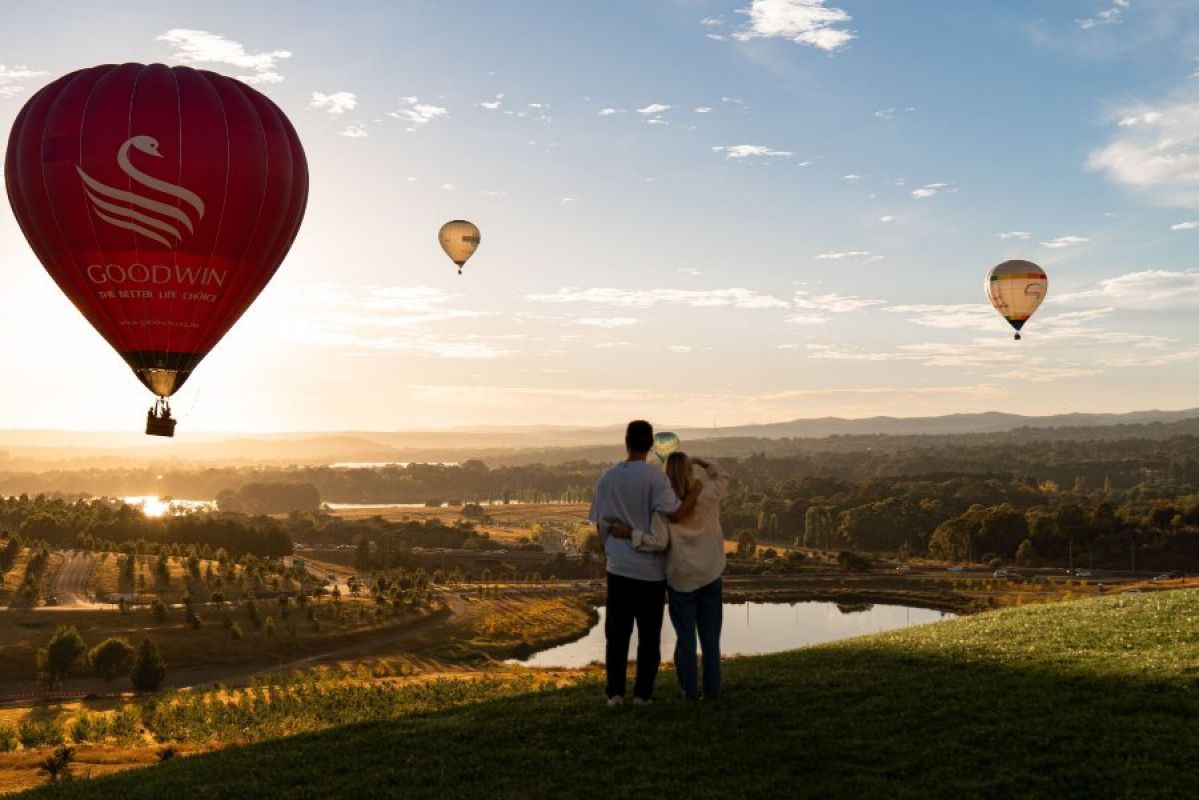 Two people with their arms around each other look out over scrivener dam from the National Arboretum, Canberra. It is sunrise and three hot air balloons can be seen in the sky