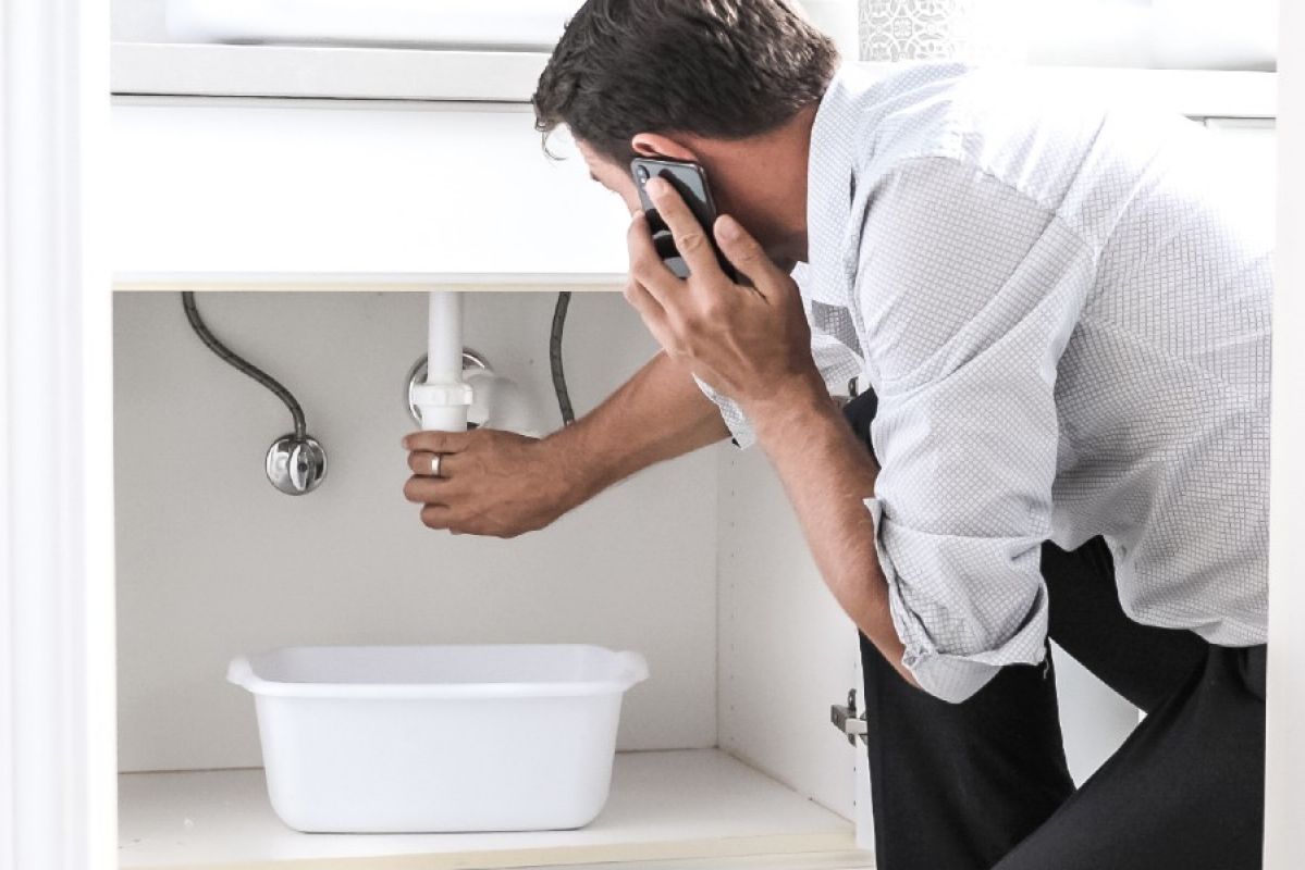 A man calling a plumber as he checks a leaking pipe under a sink