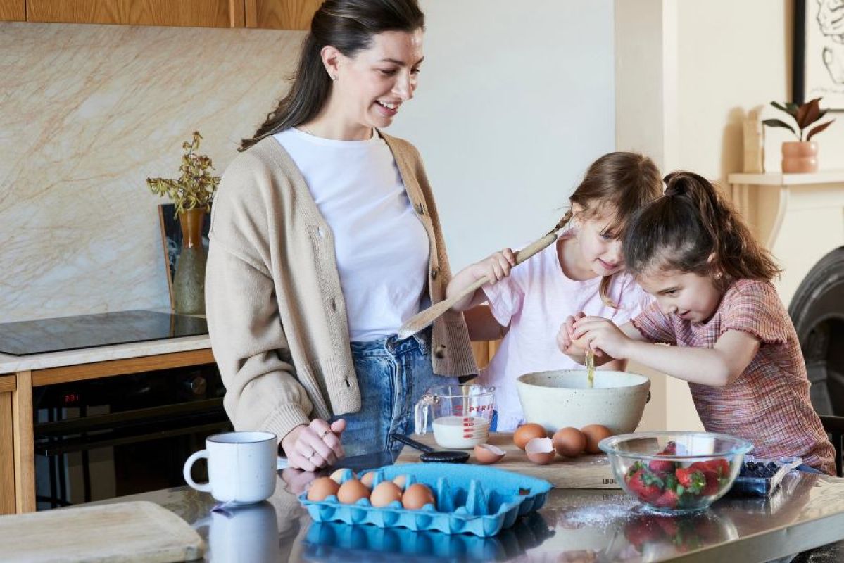 A mother and her two young daughters happily cooking at kitchen table. An induction cooktop can be seen in the background.