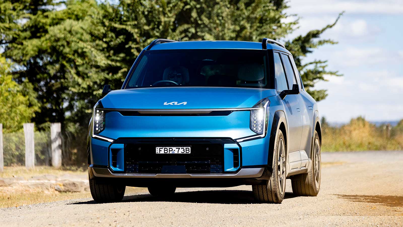 Blue Kia EV9 on a road with green trees and the blue sky behind.