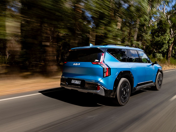A blue Kia EV9 SUV driving on the left side of a road