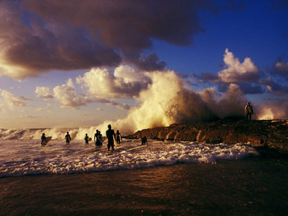 Watercolour image of surfers at a beach with waves carshing against a rock. 