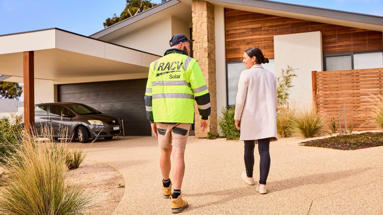 RACV Solar installer walking with a customer towards the entrance of their house, with a black car parked in the carport nearby.