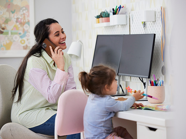 Parent laughing on the phone and young child drawing while sitting at a desk together.