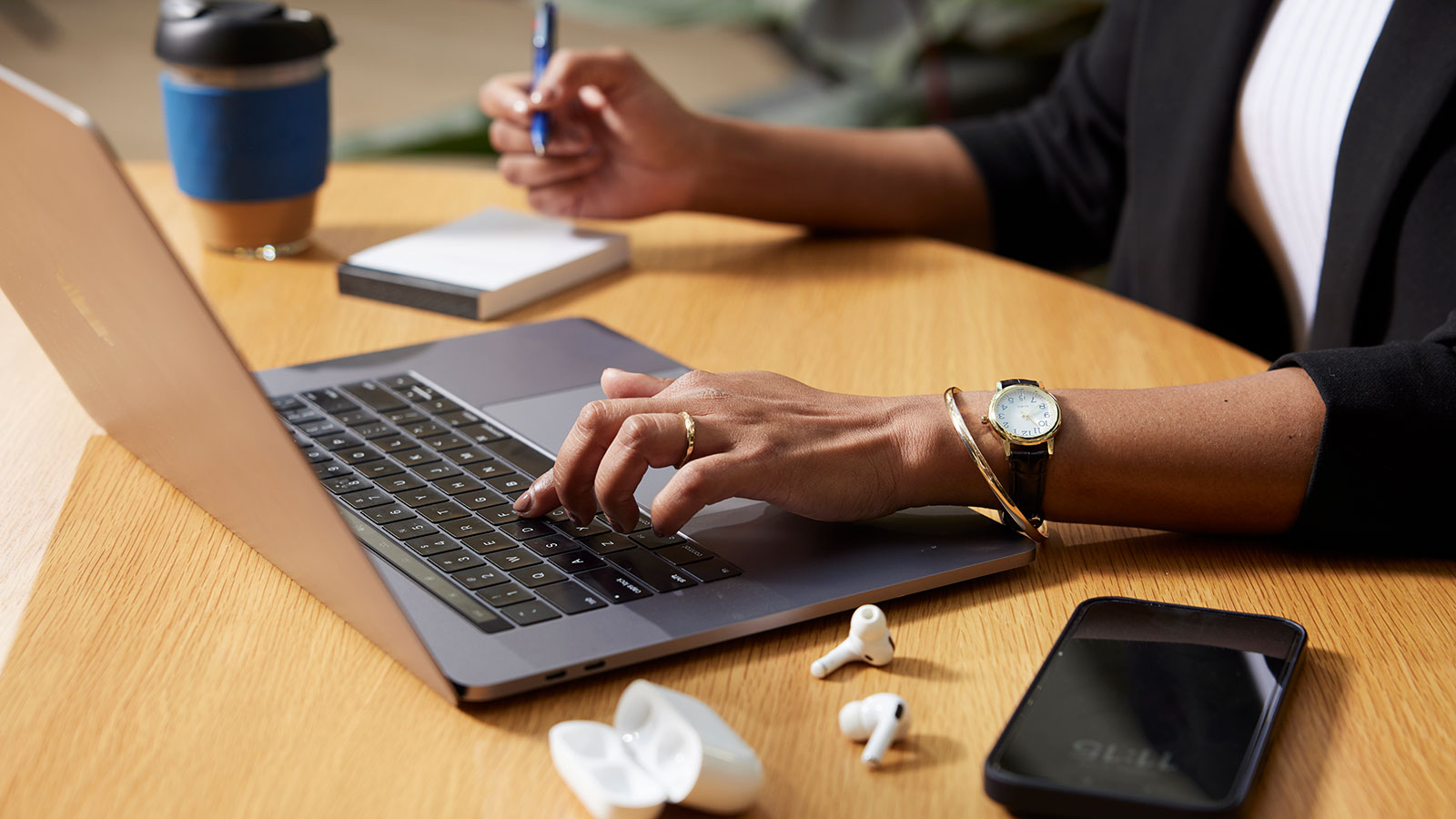 Person at a desk, using their laptop with their left hand and writing in a notebook with their right hand. Their phone, earbuds and a coffee keep cup are beside them.