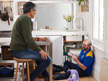 Plumber sitting on the ground fixing kitchen sink while talking to the homeowner.