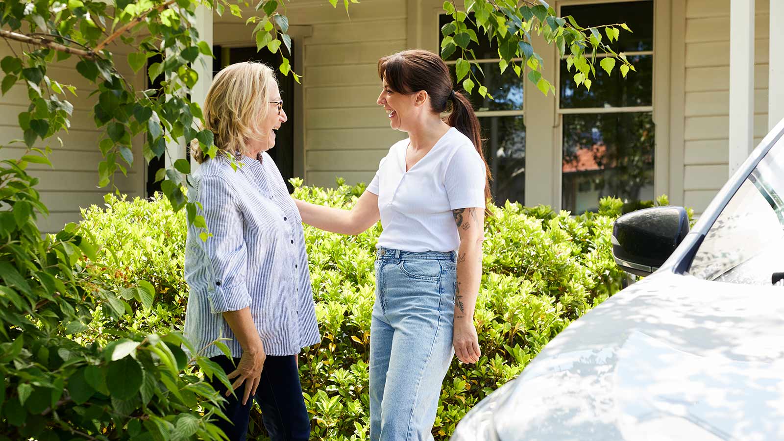 Two people smiling and greeting each other in front of a house, with a silver car beside them.
