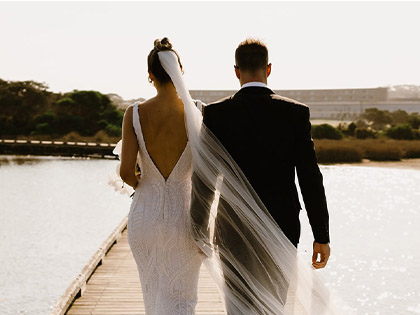 Couple walking on a pier with their backs to the camera, wearing a wedding dress and suit. 